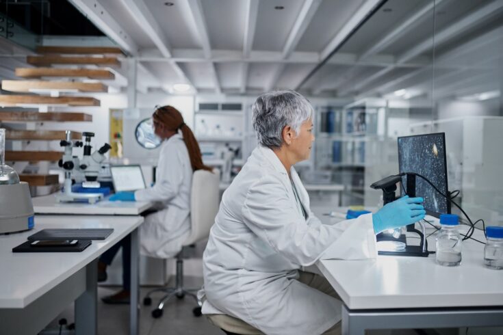 A female scientist using a computer and a digital microscope in a laboratory with her colleague in the background