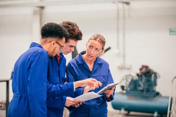 Two young men and stand in an engineering workshop with their tutor as they discuss ideas for a project. They are all wearing blue coveralls.