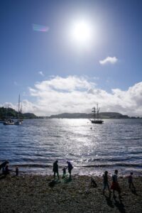 Families stand on a rocky shore, with boats floating on the sea