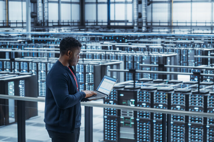 Data center engineer using laptop computer. Server farm cloud computing specialist facility with African American male system administrator working with data protection network for cyber security.