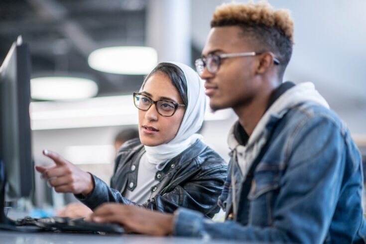 A young man and woman are sitting in a computer lab. The woman is helping with a computer project.