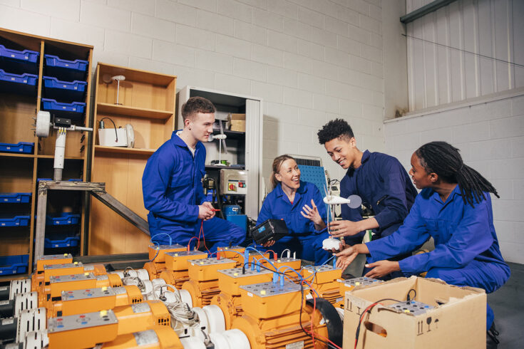 A group of engineering students and their tutor work on a group project together. They are all wearing blue coveralls and one of the students is holding a 3D wind turbine structure.