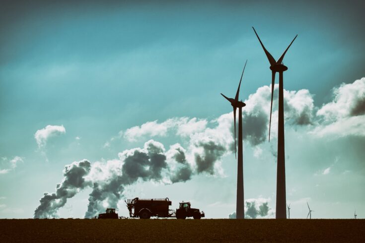 Silhouettes of two wind turbines and two tractors, pollution of coal burning power plants in the background