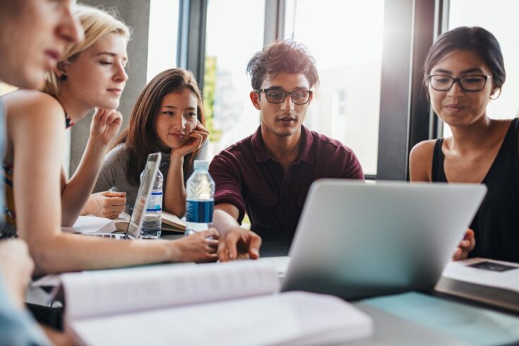 Group of students sitting at table in library and using laptop.