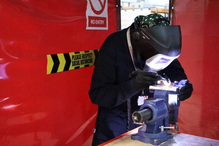 A man wearing a welding mask leans over a piece of equipment in a clamp and welds. There is light from the welding. In the background there are red safety barriers.