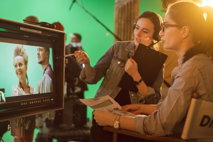 Female director in chair looking at TV display and talking with an assistant with actors and a green screen in the background
