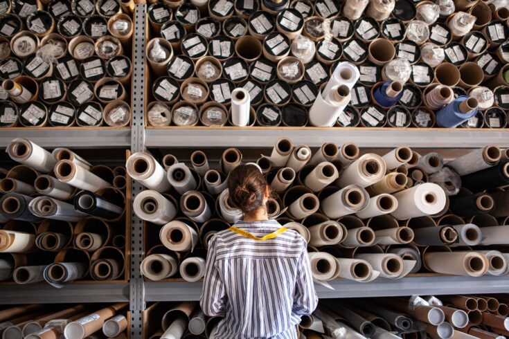 Female textile designer choosing fabric from stack of rolls inside sustainable workshop
