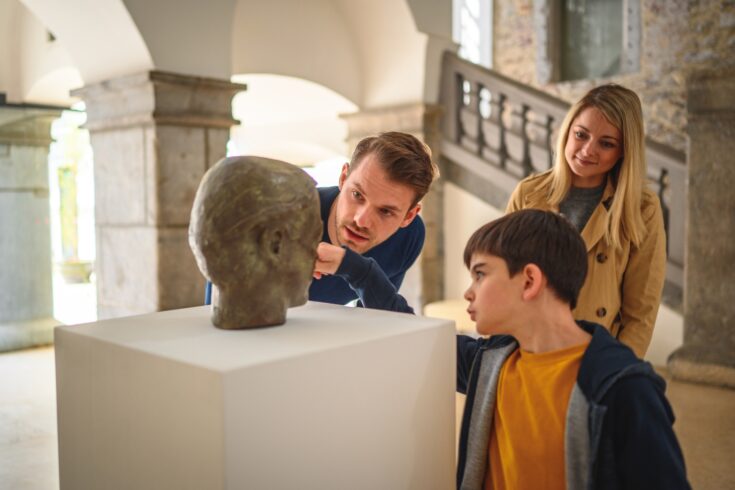 Curious boy taking a look and pointing a finger at the displayed bronze head, while his grown up siblings join him with interest.
