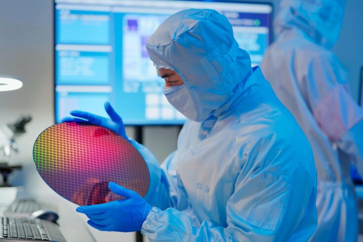 Technician in sterile coverall holds wafer that reflects many different colors with gloves and check it at semiconductor manufacturing plant
