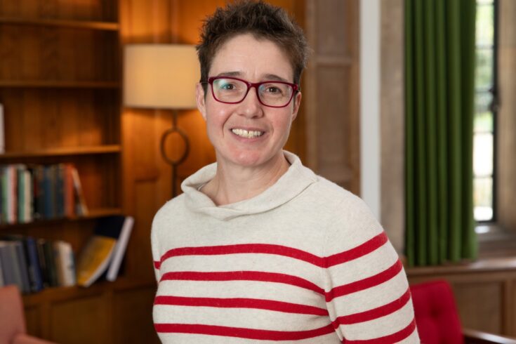 Charlotte Deane, wearing red glasses and a stripped long-sleeved t-shirt, smiling and looking into the camera