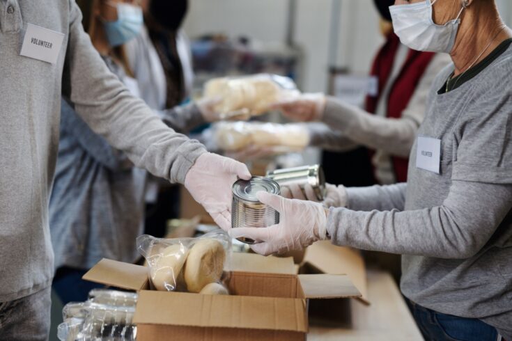 Two people dressed in grey with gloves and masks and a name tag stating 'Volunteer' holding a silver tin can at a food bank