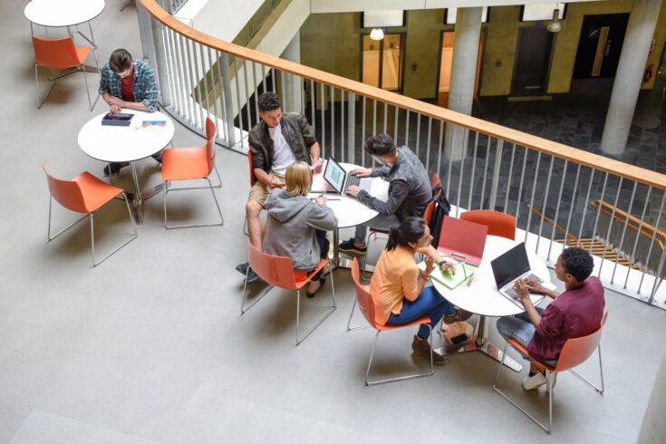 A group of students sitting at tables using laptops in a open plan space