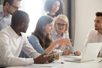 Young female mentor leader coach teaching employees group analyzing online project explaining business strategy speaking training diverse corporate team with laptop using computer at office meeting.
