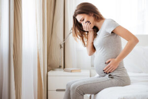 Pregnant young woman sitting on bed and feeling sick.