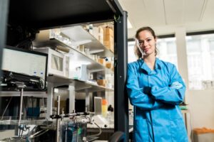 Sophie Morse wearing a blue lab coat smiling and leaning against shelf in a lab