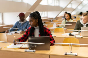 Young afro-American student is writing in the library. More students are sitting behind her.