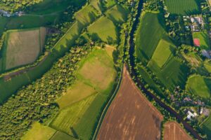 Vibrant green crops, ploughed fields and pasture, hedgerows and woodland surrounding farms and country homes beside an idyllic rural patchwork quilt landscape from high above.