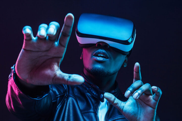 Young african man wearing virtual reality goggles with hands up, isolated on black background.