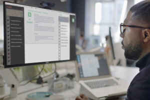 A man’s face looking into a computer screen in an open plan working office. Type is being added to the screen by an Artificial intelligence, AI, chatbot.