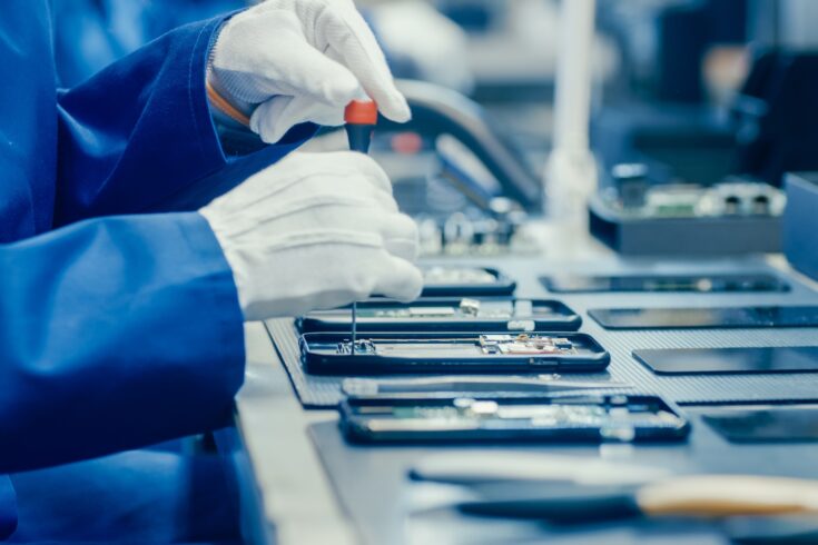 Electronics factory worker in blue work coat and gloves assembling smartphones with screwdriver