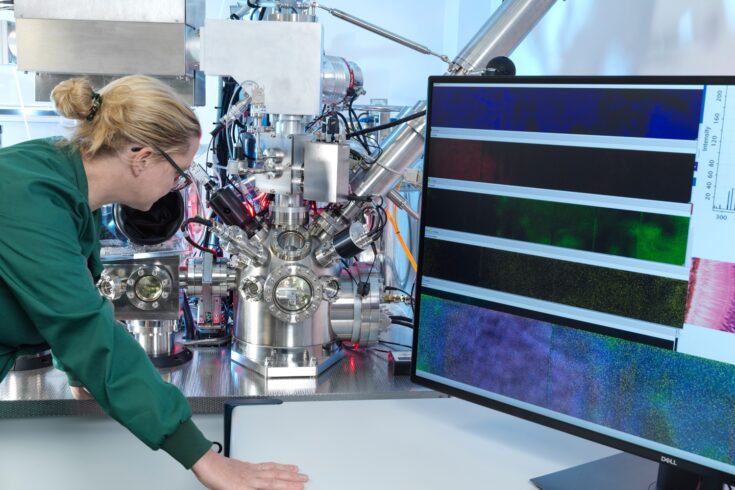Dr Felicia Green, a lady wearing a green lab coat, looking at a silver dome with silver and black pipes coming out of it, which is the stigmatic imaging secondary ion mass spectrometry (SIMS) instrument.