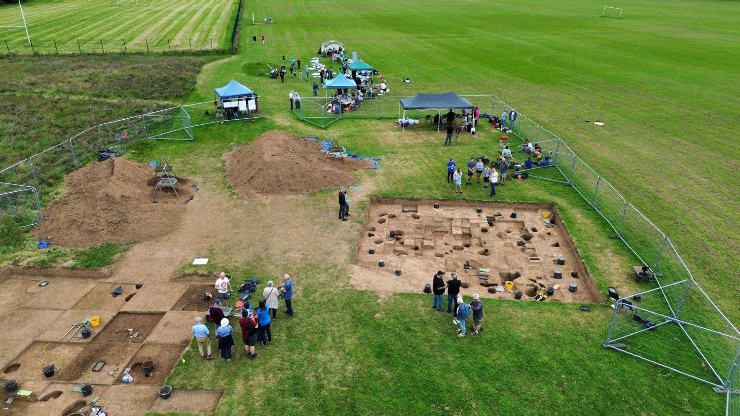 An aerial view of an archaeological excavation site in the Caerau Hillfort with several square pits, people working, and tents set up for shade or equipment storage. The site is in a grassy field with some equipment and dirt piles nearby.