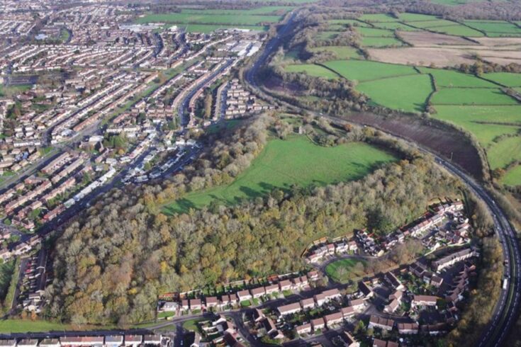 An aerial view of Caerau Hillfort in Wales, showing residential housing, green spaces, and woodland, with a curved road or railway line running through the area
