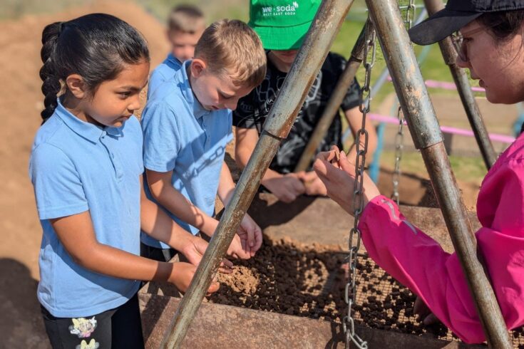 Children in blue shirts are participating in an archaeological activity, gathered around a metal frame with a sifting tray filled with dirt. An adult in a green hat and another person in a pink jacket are assisting them. The background shows an outdoor setting with greenery and dirt paths, indicating a hands-on heritage learning experience