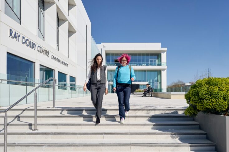 Two people walking down steps outside the Ray Dolby Centre building in Cambridge