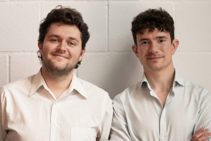 Two white men wearing white shirts smiling at the camera, against a white painted wall