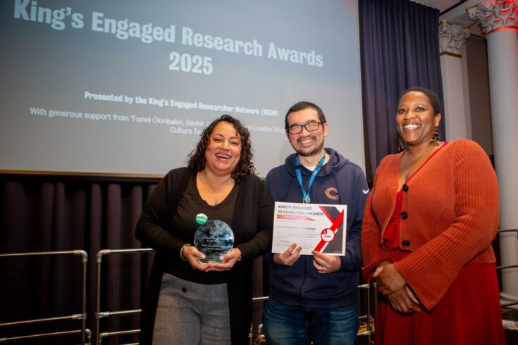 Three people standing and smiling, the person on the left is holding an award and the person in the middle is holding the award certificate
