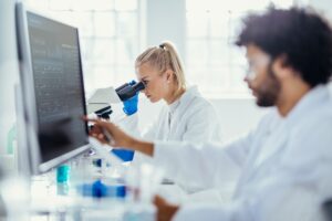 Two people wearing white lab coats, concentrate on analyzing specimen under a microscope, in a bright laboratory setting.