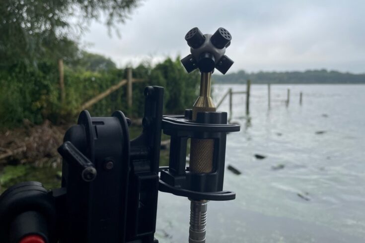 Microphone on a tripod recording beside a lakeshore with audio gear on the ground