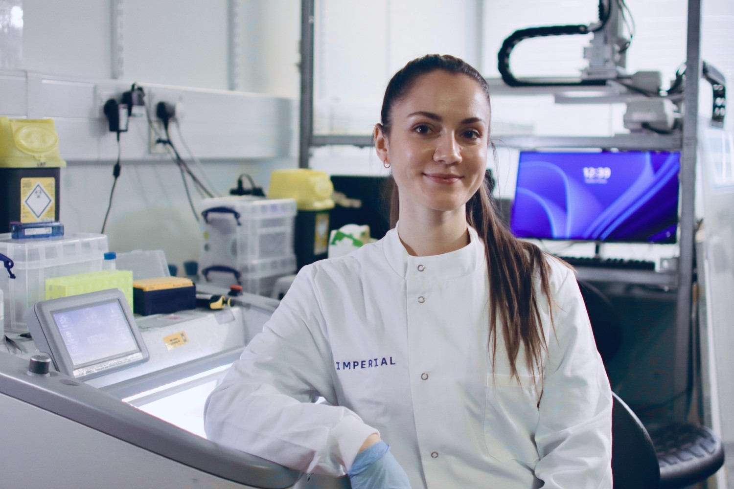 A young woman wearing a lab coat with 'Imperial' written on it, sitting in a lab. 
