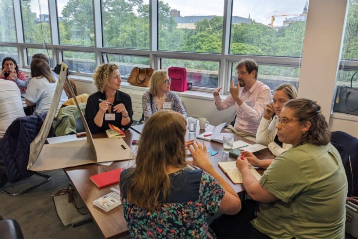 A group of adults in discussion in a room overlooking a park with paper and a flipchart on the table in front of them