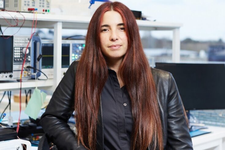 A person with long, straight dark hair sits in a modern electronics laboratory. The individual is wearing a black outfit with a dark leather jacket and is seated in front of a workstation filled with electronic testing equipment, cables, and monitors. Shelving units behind the person hold various instruments, and large windows in the background show an outdoor view with a bright sky.