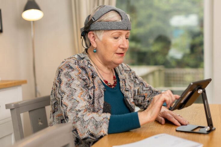 An older adult seated at a table wearing a neuroscience headset using a tablet mounted on a stand. The person is wearing headphones and a patterned cardigan, and is tapping the tablet screen with one hand.
