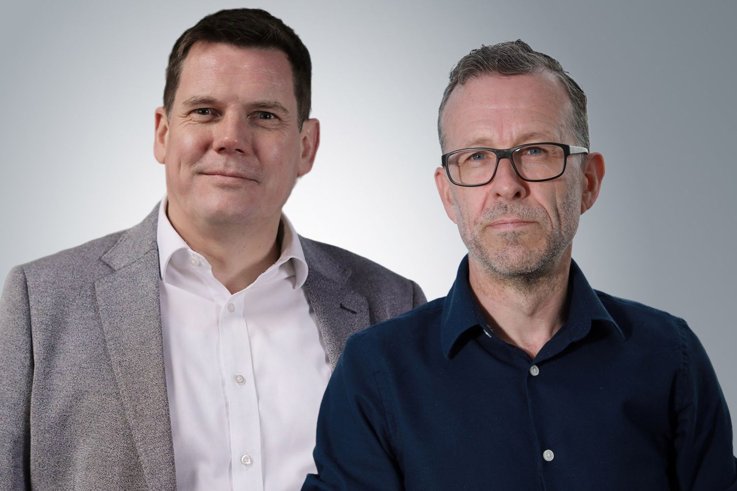 Two adults standing side by side against a plain gradient background. One is wearing a light-coloured blazer over a buttoned shirt, and the other is wearing a dark long-sleeved shirt and glasses.
