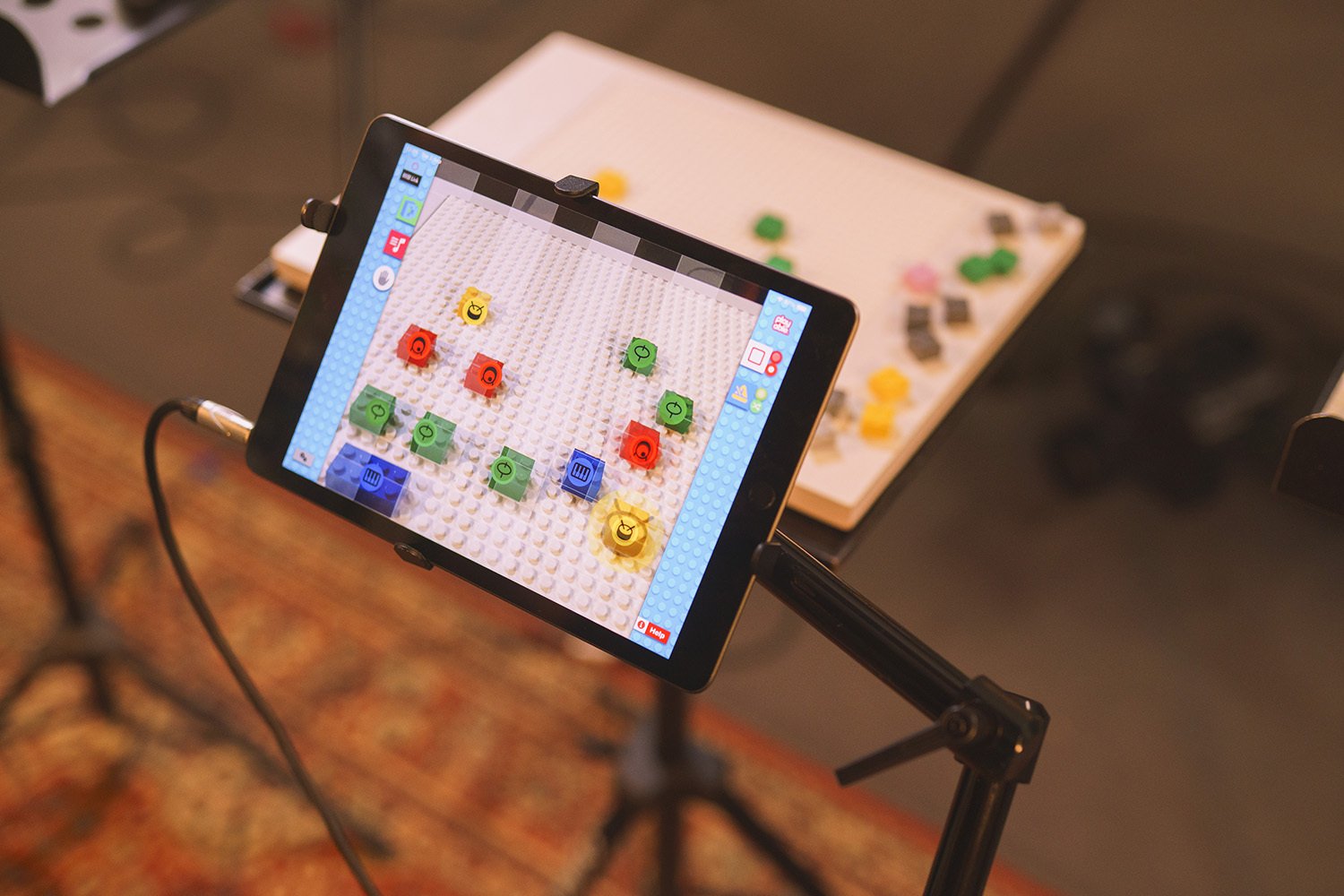 A screen on a tablet device displaying LEGO bricks. A physical LEGO base plate with LEGO bricks can be seen on a table behind it.