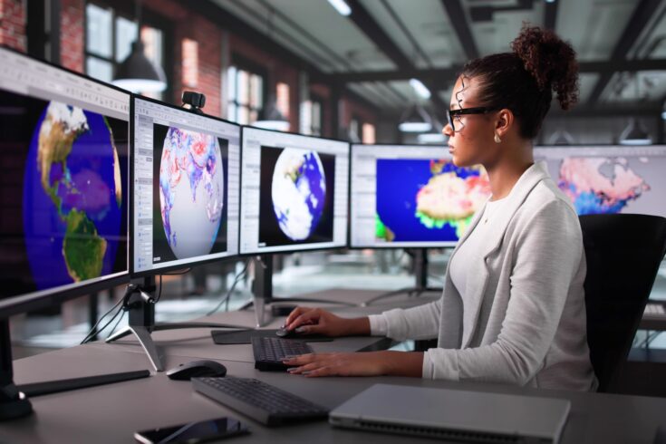 A woman sits at an office desk, working on multiple large computer monitors displaying global maps and satellite imagery.
