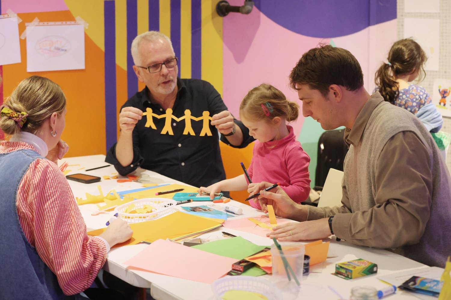 A man is holding a paper chain of connected people, next to him a little girl is drawing and is accompanied by her parents