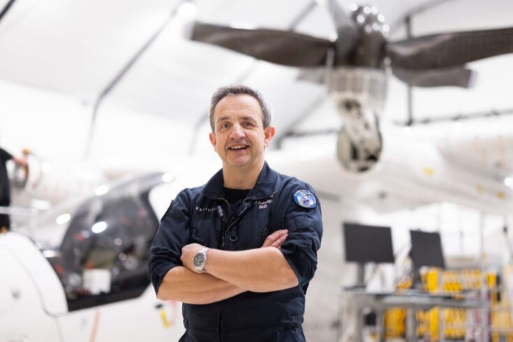 Person in a dark flight suit standing with arms folded in an aircraft hangar, with a helicopter and rotor blades visible in the background.