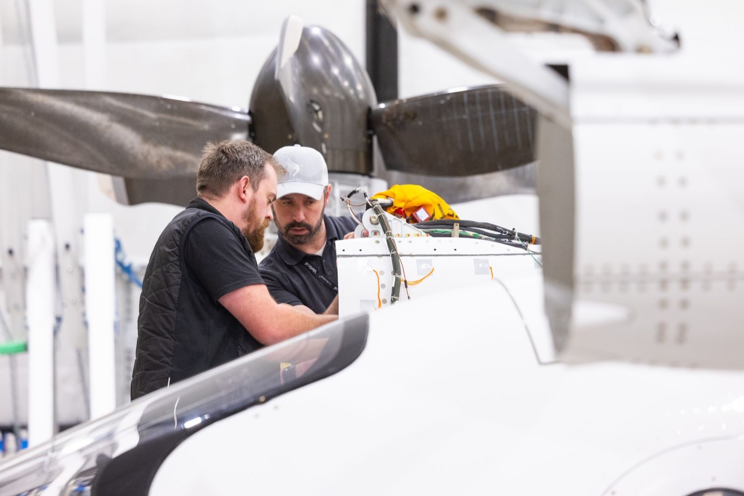 Two engineers working on aircraft equipment inside a hangar, with helicopter rotor blades visible in the background.
