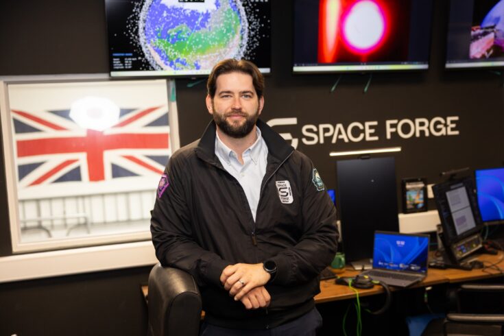 Person wearing a Space Forge jacket stands beside a workstation in a mission control-style room. Multiple screens behind display satellite and space-related visualisations, while the Space Forge logo, computer monitors and a UK flag are visible in the background.
